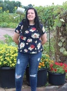 Lizzy is wearing a blue shirt with white and pink flowers., along with wearing blue jeans. Lizzy is standing in front of red, orange and yellow flowers and a green vine.