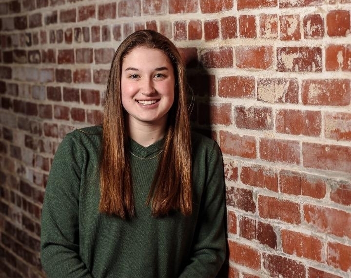 Emma is wearing a long sleeve green sweater. She is leaning up against a brown and white brick wall.