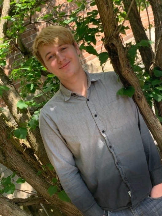 Wiant is wearing a grey dress shirt, standing in front of a tree, with one hand in his pants pocket. The tree is brown and green