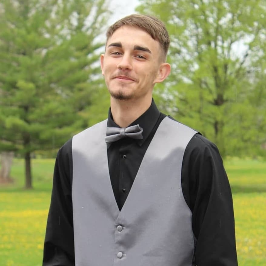 Nathan is wearing a black long sleeve dress shirt with a grey vest. He is standing in yard that is fully green with trees in the background.