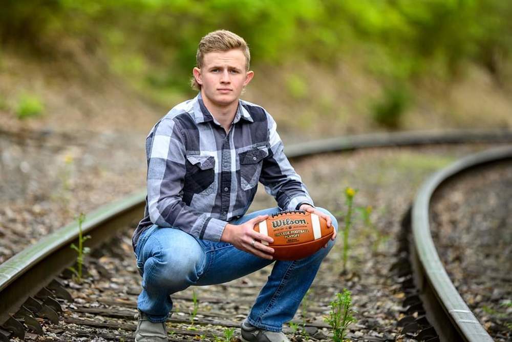 Mack is squating down hold a football with in a set of railroad tracks. He is wearing a black/grey/white long sleeve shirt and blue jeans.