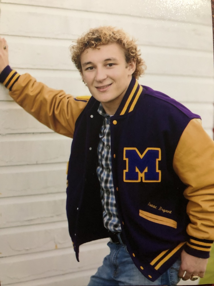 Isaac is wearing his purple and glod Varsity jacket. He is also wearing a blue and black plaid shirt and blue jeans. Issac is leaning up the side of a white house.