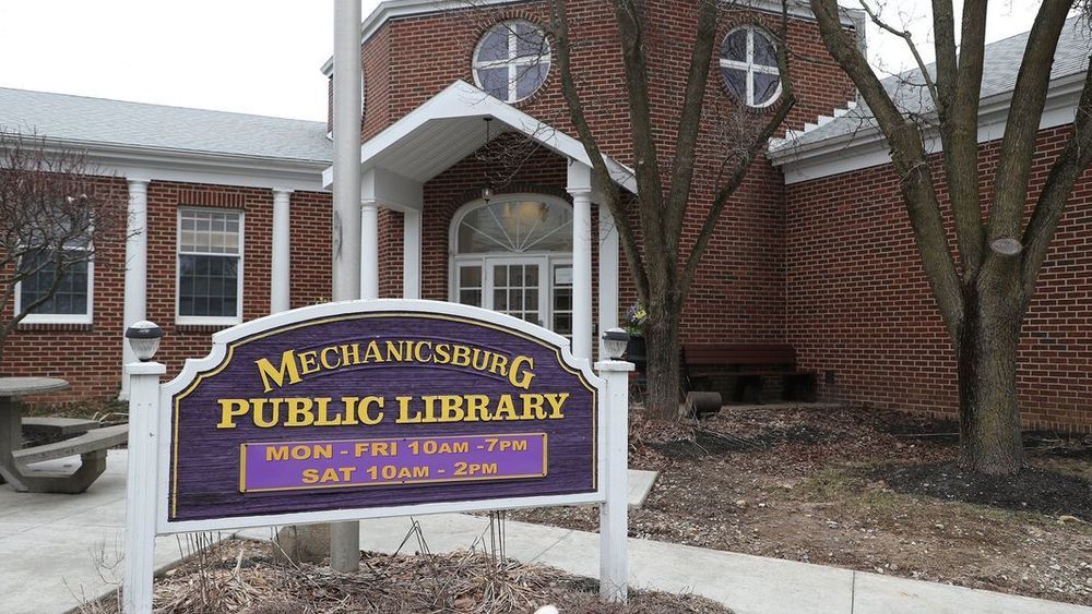 Mechanicsburg Puplic Library ( in yellow) on a purple and white sign in front of the Library. The library is brown and white. The trees are bare, it looks like its the end of winter.
