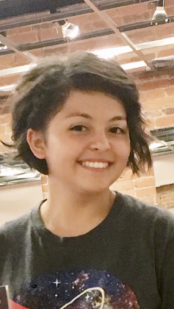 Karla is wearing a black shirt with some blue, red and gold on it. She standing in front of something brick. 