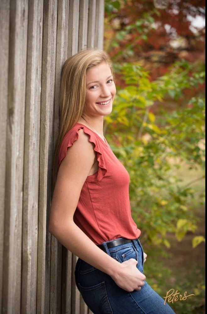 Kasey is wearing a pick top with blue jeans. She is leaning up against a wooden fence with her hands in her pockets. behind her is green, brown trees. 