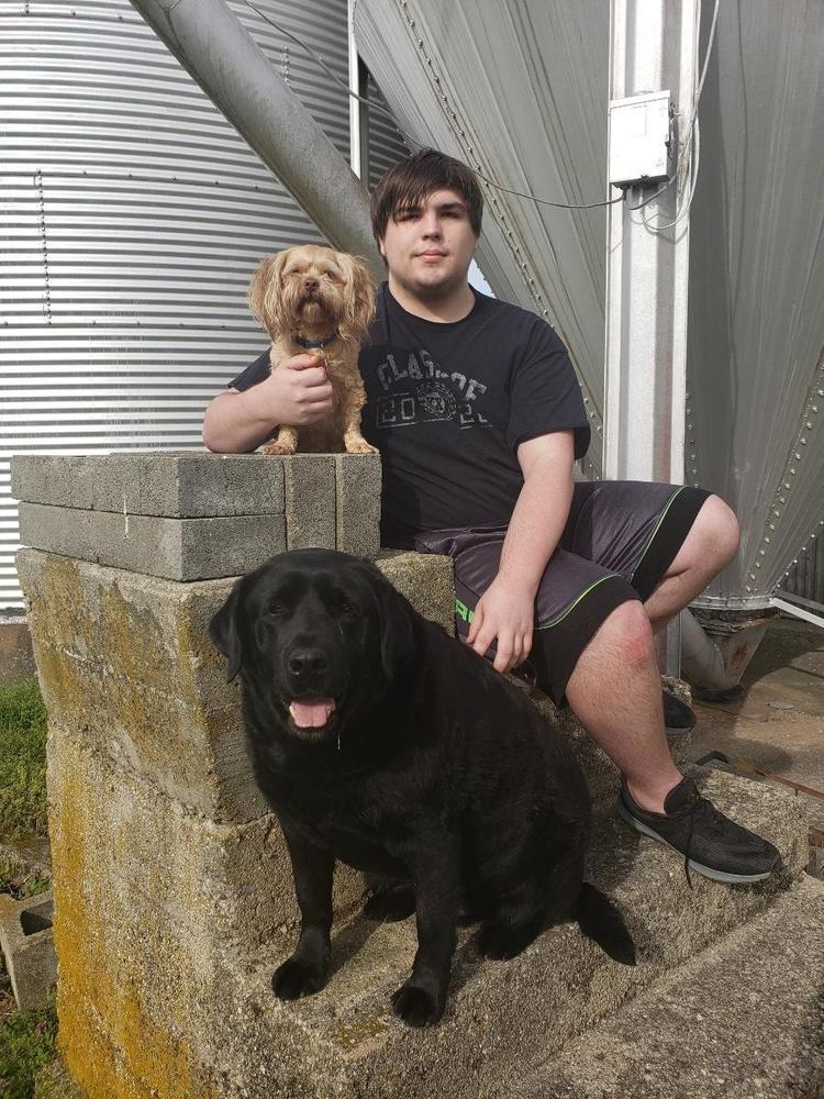 Izzy is wearing a black shirt and shorts. He is sitting beside his dogs on a step on a porch. One of Izzy dogs are black and the other is brown.