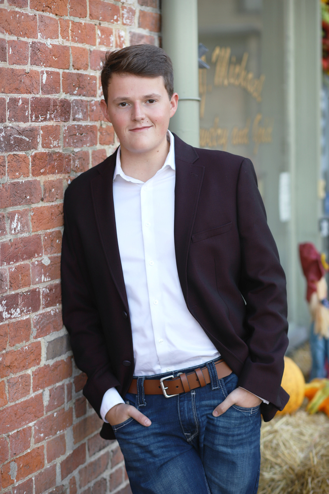 Ross is wearing a brown suit jacket with a white shirt, blue jeans and brown belt. Ross is leaning up against a white and brown brick wall.