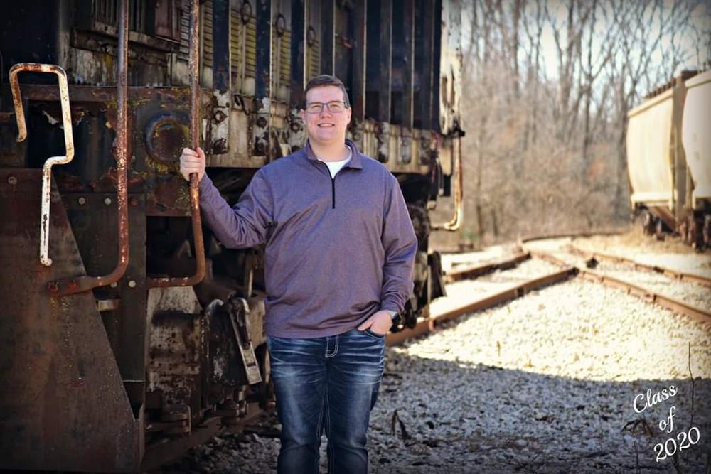 Evan is wearing a purple long sleeve quarter zip, alond with blue jeans. Evan is holding on to a railing of a train that is stopped on the railroad tracks. He is standing in the gravel and in the background you can see trees that have lost their leaves and another train.