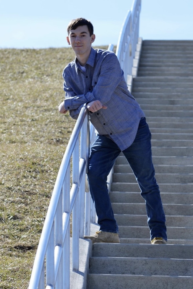Trenton is wearing a long sleeve blue shirt and blue jeans. He is leaning up against a white railing. The railing is attached to grey staris, which Trenton is standing on. 