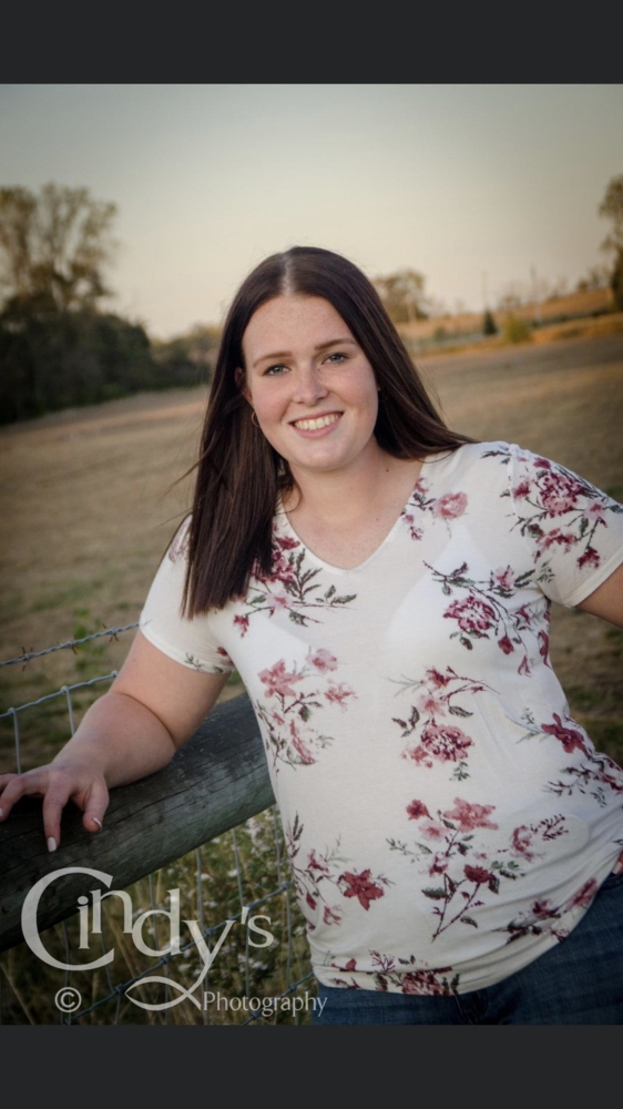 Morgan is wearing a white top with pink flowers with blue jeans. Morgan is leaning up against a wood fence. with the background being a field.