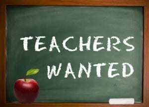 Teachers Wanted written in white on a green chalkboard. The outline of the chalkboard is brown. Also on the chalkboard is a red apple with a green stem on the left and on the right side is a piece of white chalk