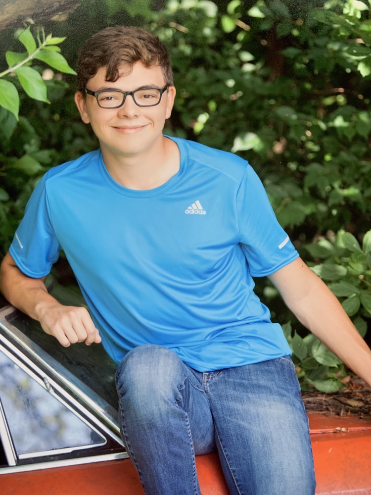 Trent is wearing a blue tshirt and blue jeans. Trent is sitting on an orange car. That is sitting in front of a green tree. 