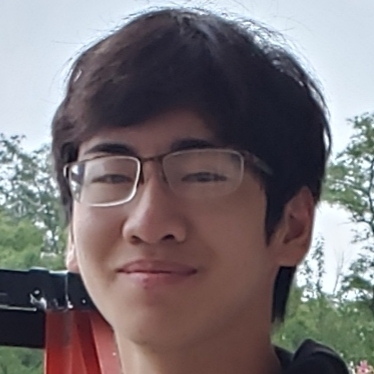 Andrew is wearing glasses with a white frame, a black shirt and is standing outside with green treas in the background.