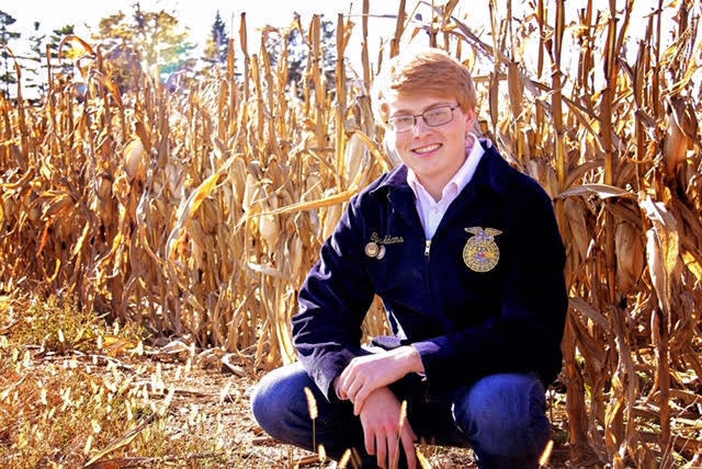 Tyson is squating down in a corn field. He is wearing his FFA Jacket with blue jeans. Tyson is also wearing glasses with white frames.