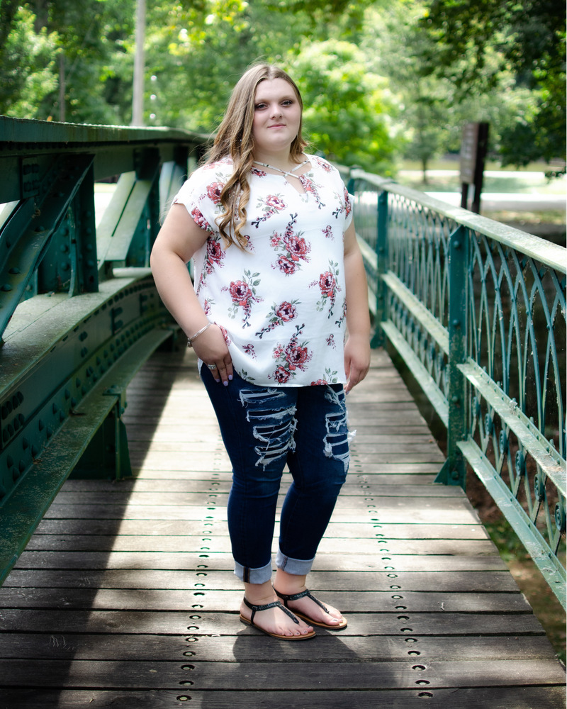 Madison is wearing a white shirt with pink flowers and blue jeans. Madison is standing on a wooden bridge that has green railing. There are also green trees in the background.