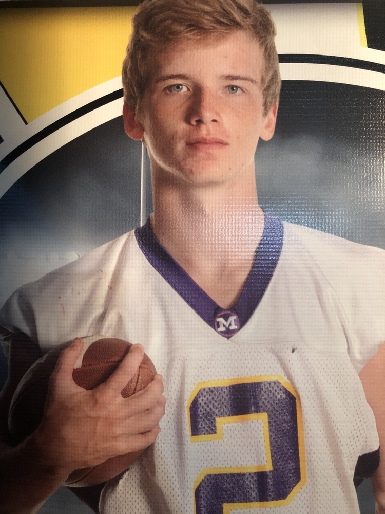 Kyle, is wearing his football jersery that is white, purple and gold. He is holding a football. The background is grey, white and yellow