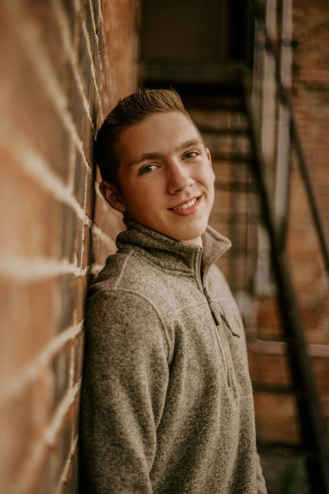 Wyatt is wearing a brown long sleeve.Leaning up against a brown brick wall. With a black stairway behind him.