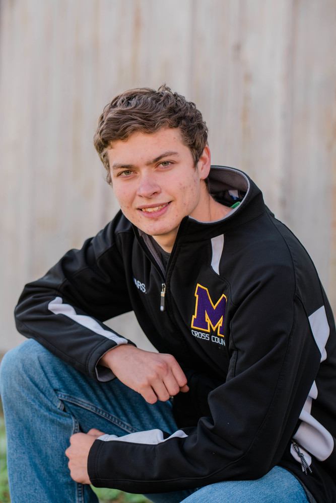 Jippy, JP is wearing a black and grey Mechanicsburg windbreaker jackte. The jacked also the M on it . He is kneeling down in front of a grey/white fence.