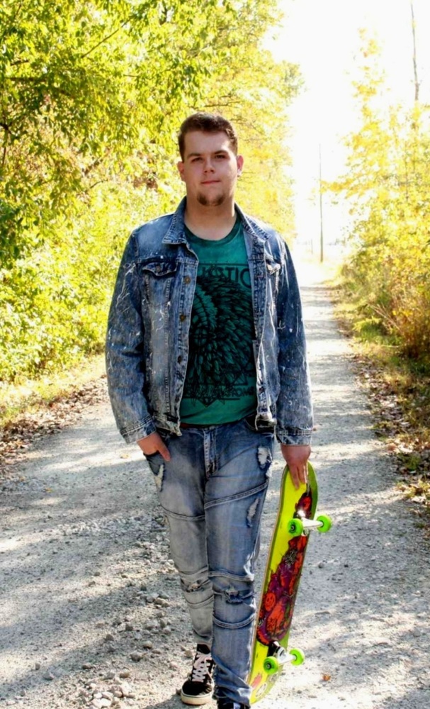 Chandler is wearing a blue jean jacket and pants. He is wearing a green shirt with black. He is holding a neon yelloow and red skateboard. He is standing on a gravel path with is tree lines.