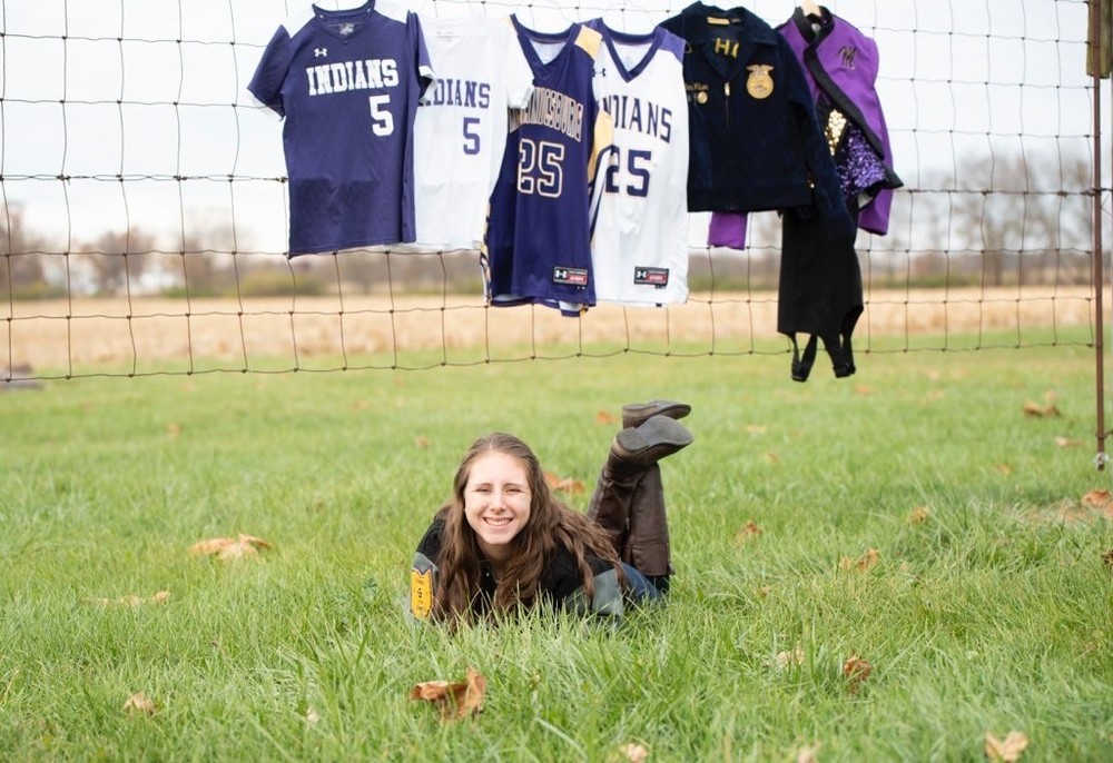 Emma is wearing black shirt and blue jeans. she laying in the grass. Behind her is a fence that is holding her jersey's from various sports. They are purple, white and yellow. Also hanging is her FFA jacket and band uniform.
