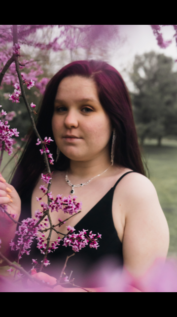 Lauren  Is wearing a a black tank top. She is standing with a purple tree.  In the background are other trees that are green. 