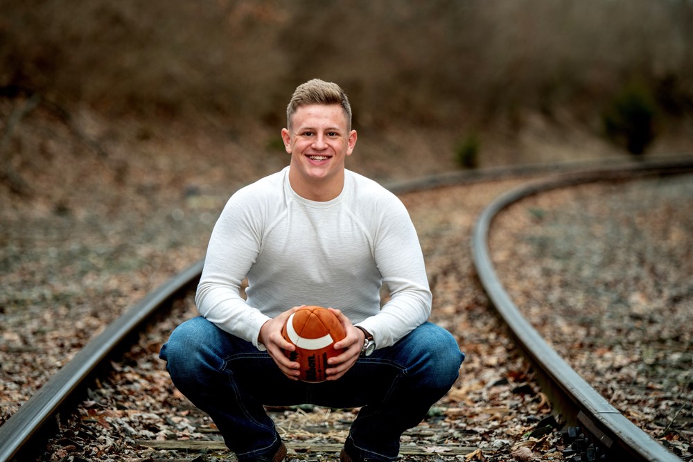 Kix is wearing a white long sleeve shirt with blue jeans. He squating, holding a football with in railroad tracks that is covered in falling leaves.