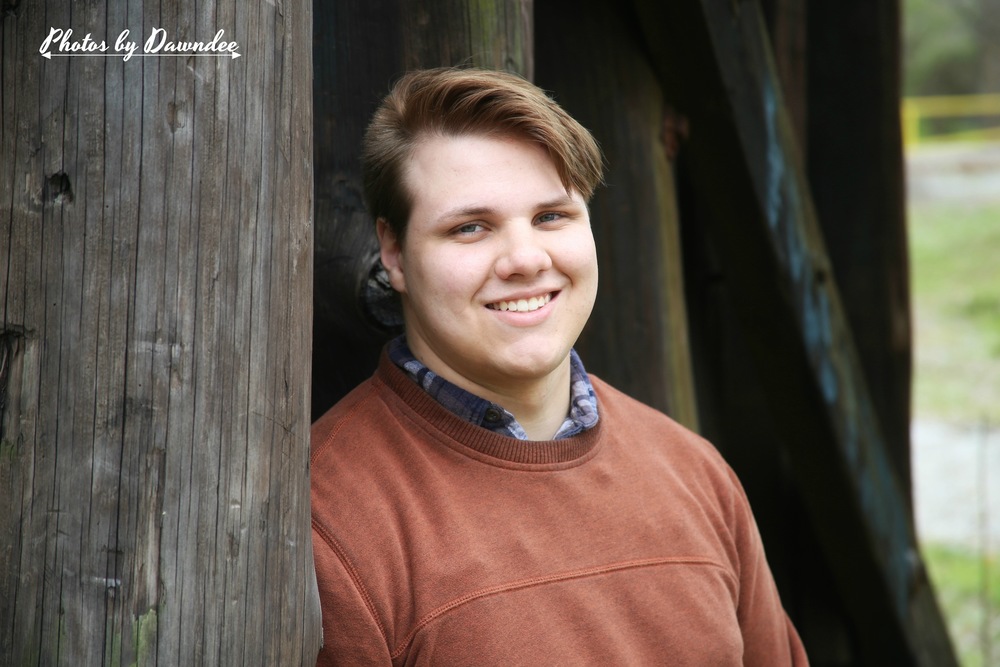 Erick is wearing a orange long sleeve sweater and blue colored shirt underneath. He is standing in between what looks like thick trees.