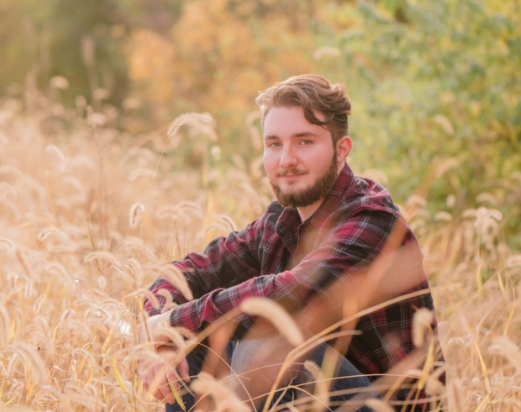 Sam is wearing a red and black plaid long sleeve shirt and blue jeans. Sam is sitting in a field. The field is turning colors.
