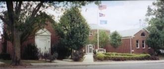 Picture of the Mechanicsburg library. The building is browqn with white doors and windows. In front of the building stands a flag pole with the flags flying. There are green trees standing in various places in front of the building