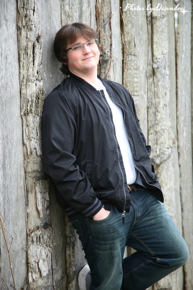 Ethan is wearing a black jacket, white tshirt, blue jeans. He is leaning up a fence with one foot proped up behind him on the fence.
