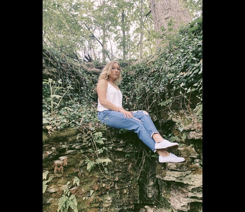 Skyler is wearing a white top, blue jeans, and white pants. She is sittin on a rock in the forrest and it very green.