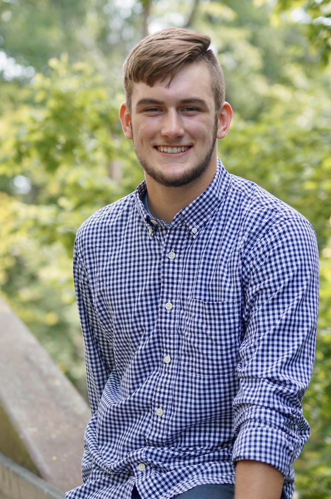 Logan is wearing a white and blue long sleeve shirt. Logan is sitting a wooden bridge with green trees in the background. 