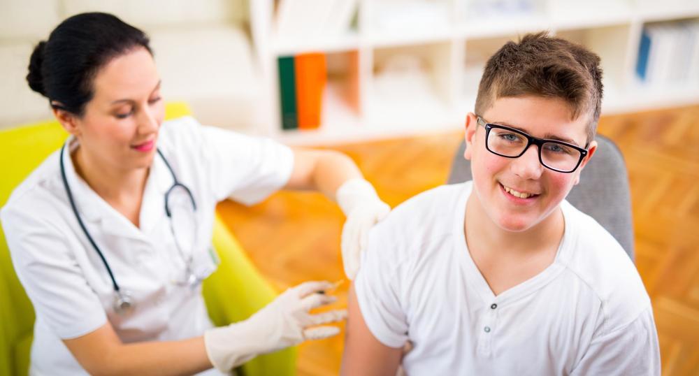 Picture of a nurse dressed in white. She is wearing white gloves. She is giving a male student a shot. The student is wearing a white t-shirt. The nurse is sitting in a neon green chair. The floor is orange. 
