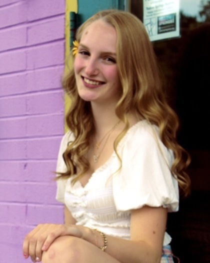 Ava is kneeling in front a black and purple wall. Ava is wearing a white top.