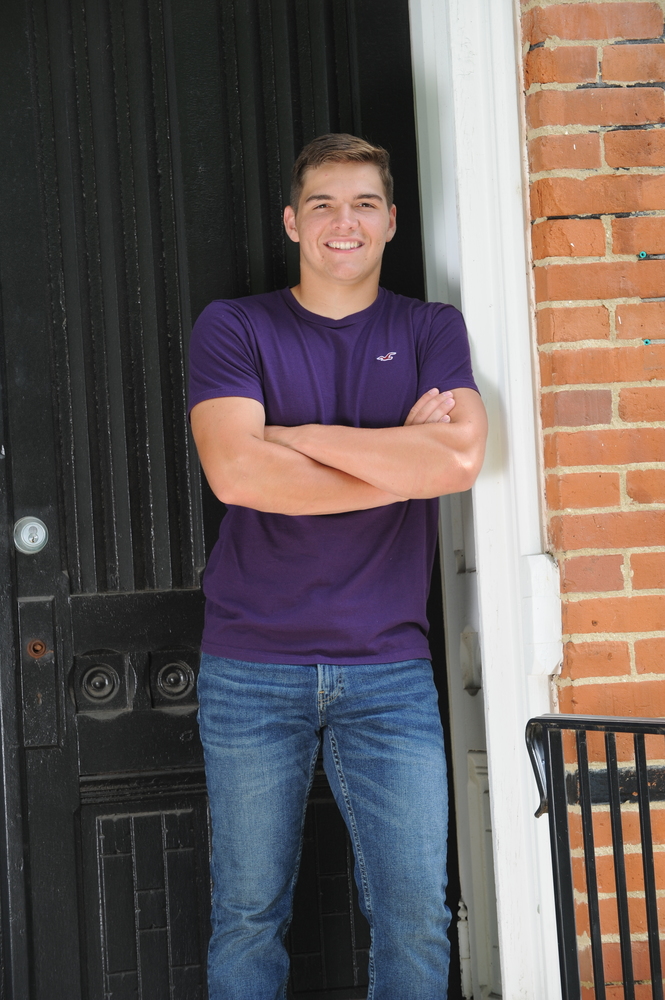 Noah is wearing a purple shirt and blue jeans. Noah is standing front a black door. Leaning up against a white frame of a brown brick building.