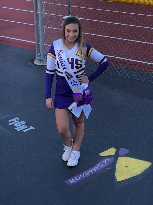 Kaylee is wearing her cheerleading uniform. Her uniform is purple, white and yellow. Wearing a Senior sash. He is standing in front of the HS track.