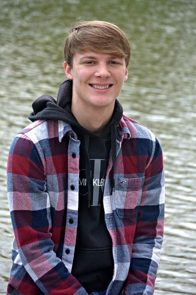 JD is wearing a blue, red, white jlong sleeve. under that he is wearing a black hoodie. JD is sitting/standing in front of a body of water.