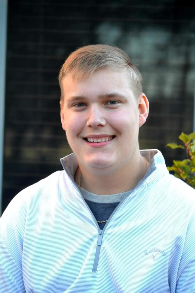 Trent is wearing a white jacket over top of a blue/grey shirt. The background is blurred out. 