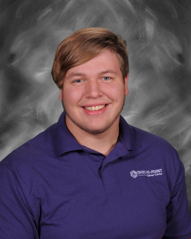 Tryston is wearing a white polo shirt. he is standing/sitting in front of a grey/black background.