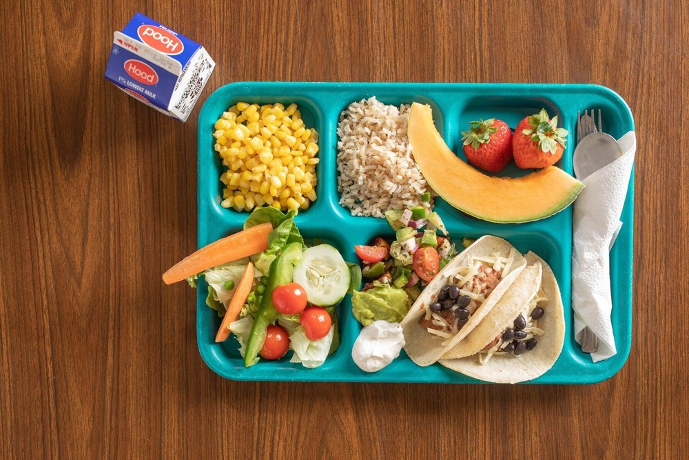 Pictue of a blue school lunch tray, with various foods on it.  Corn, rice, fruit, tacos, silverware. also picuted is a school milk