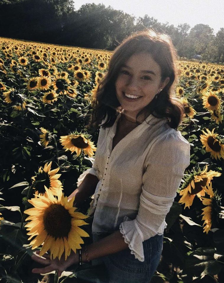 Kerrigan is wearing a white long sleeve top with blue jeans. Kerrigan is holding a sunflower standing in a sunflower field. 