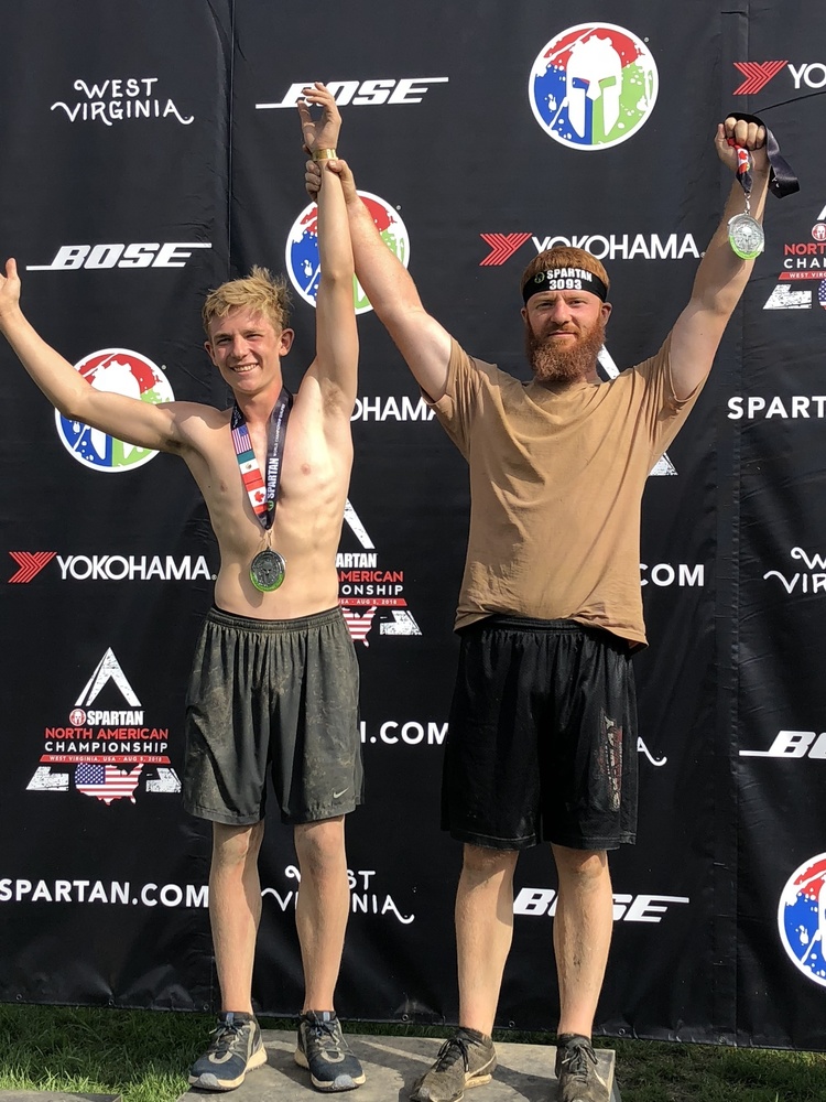 Dakota is wearing grey shorts with a medal around his neck. He is standing with another gentalman whom is wearing black shorts and a brown shirt. They are standing in front of a black back drop that has the words BOSE, Spartan and West Virginia in white on the backdrop.