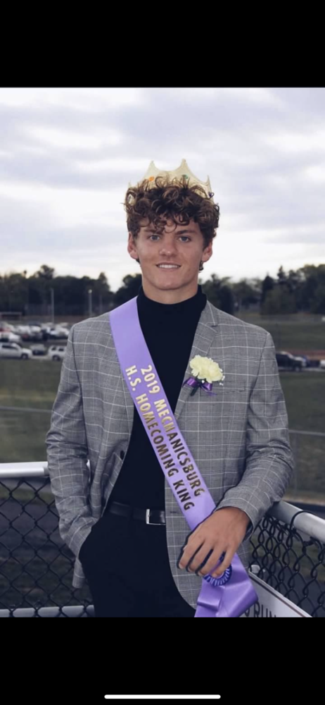 Butsch is wearing a black shirt, black pants and a grey suit jacket. Bradley is also wearing a purple sash, and gold crown. He is leaning up against a silver fence with a field and cars in the background.