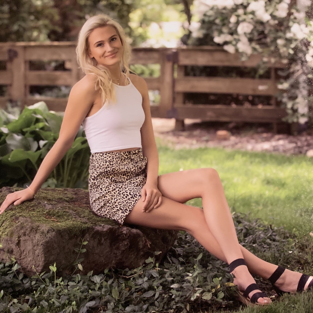 Macie is wearing a white tank top with a brown leopard skirt, with black sandals. She is sitting a brown rock with green grass in the backgroun. Also in the background is a brown fence and white flowers