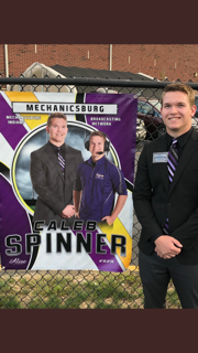 Caleb is wearing a black suit., with a purple tie. Caleb is standing in front of a fence. The fence is holding his senior banner. The banner says Caleb Spinner at the bottem. At the top is Mechanicsburg. Caleb's banner has a picture of Caleb in a black suit and another wearing a purple shirt and an announcer headset.