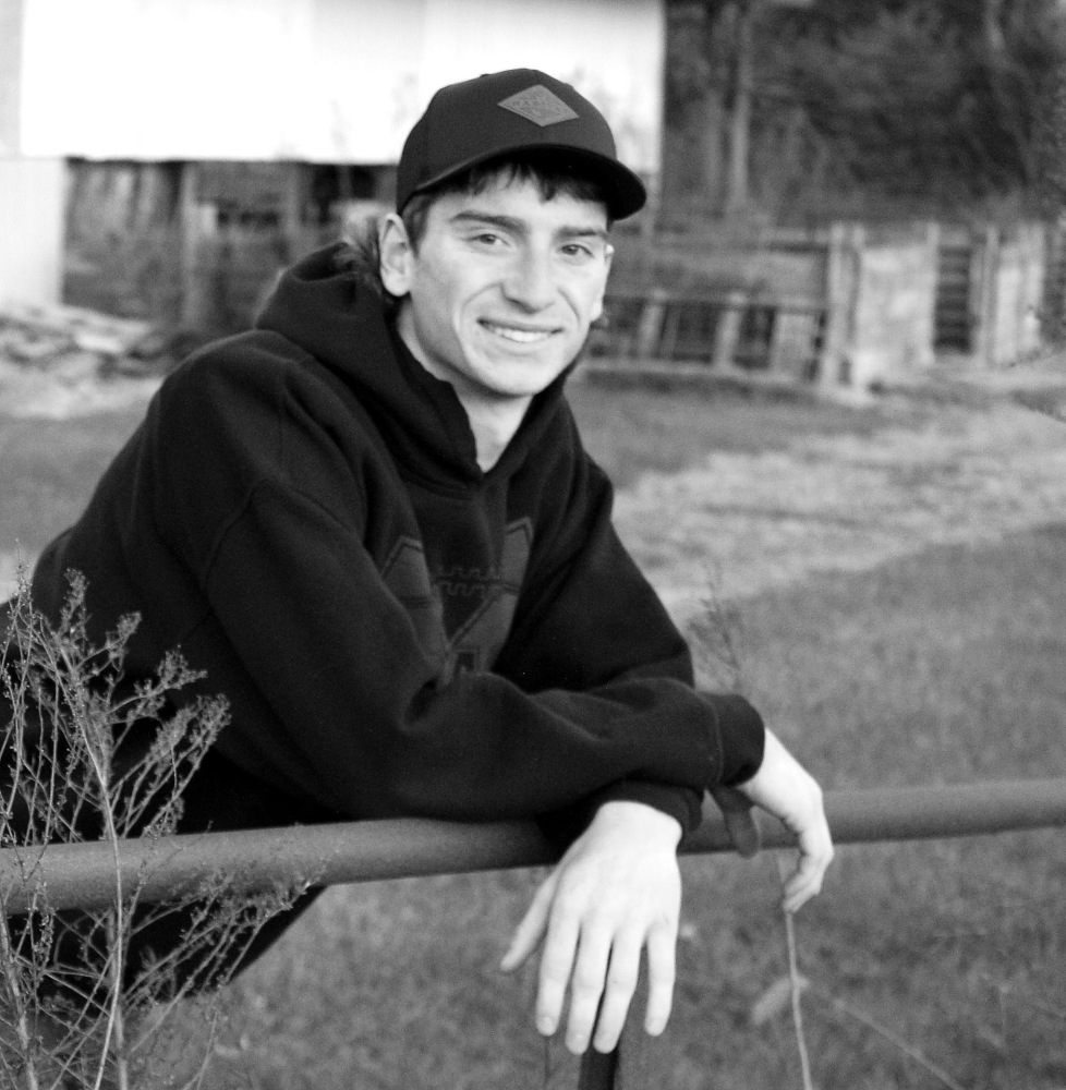 Colin's picture is black and white. Colin is wearing a hoody and a ball cap. Colin leaning up againts a gate , looks like on a farm