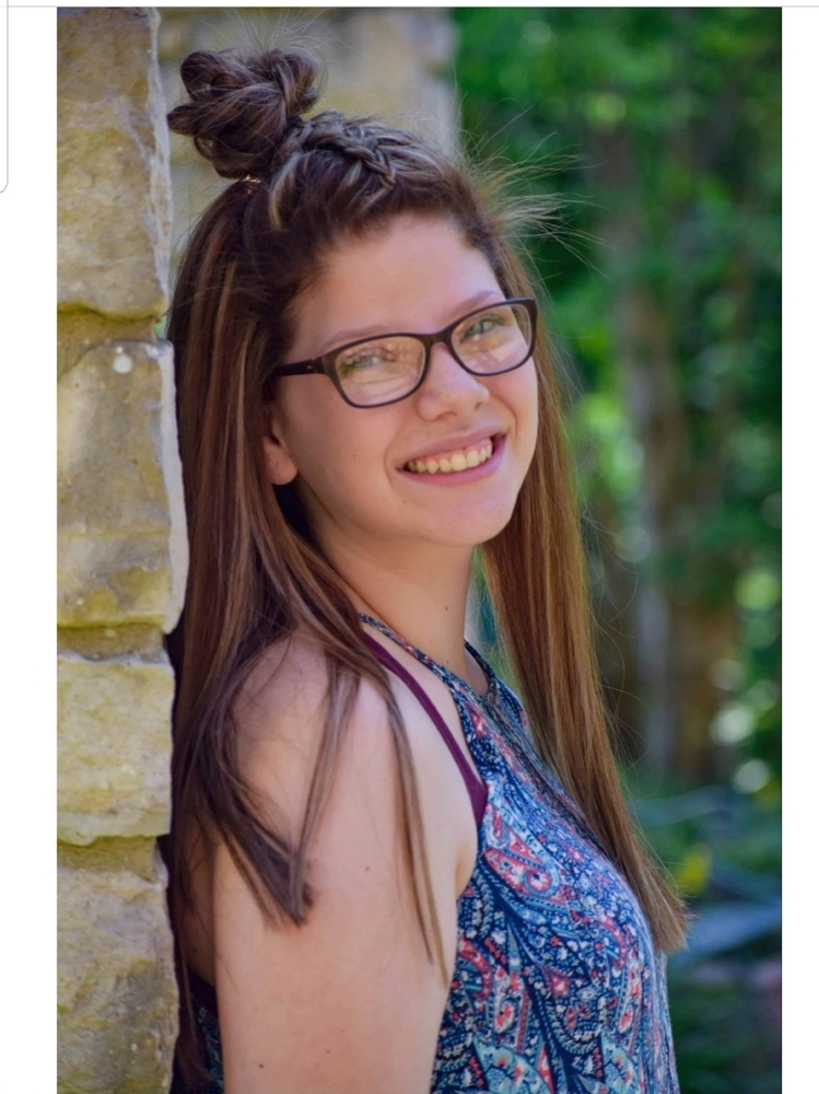 Donna is wearing a blue top. She leaning up against a brick wall with trees in the background. Donna has half her hair pulled up and is wearing black glasses.