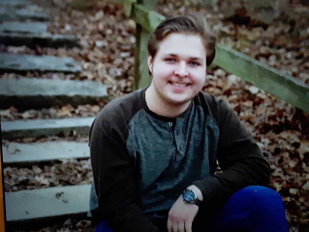 Tristan is wearing a brown and grey long sleve shirt with blue jeans. He is sitting on steps during fall time and leaves have fallin.