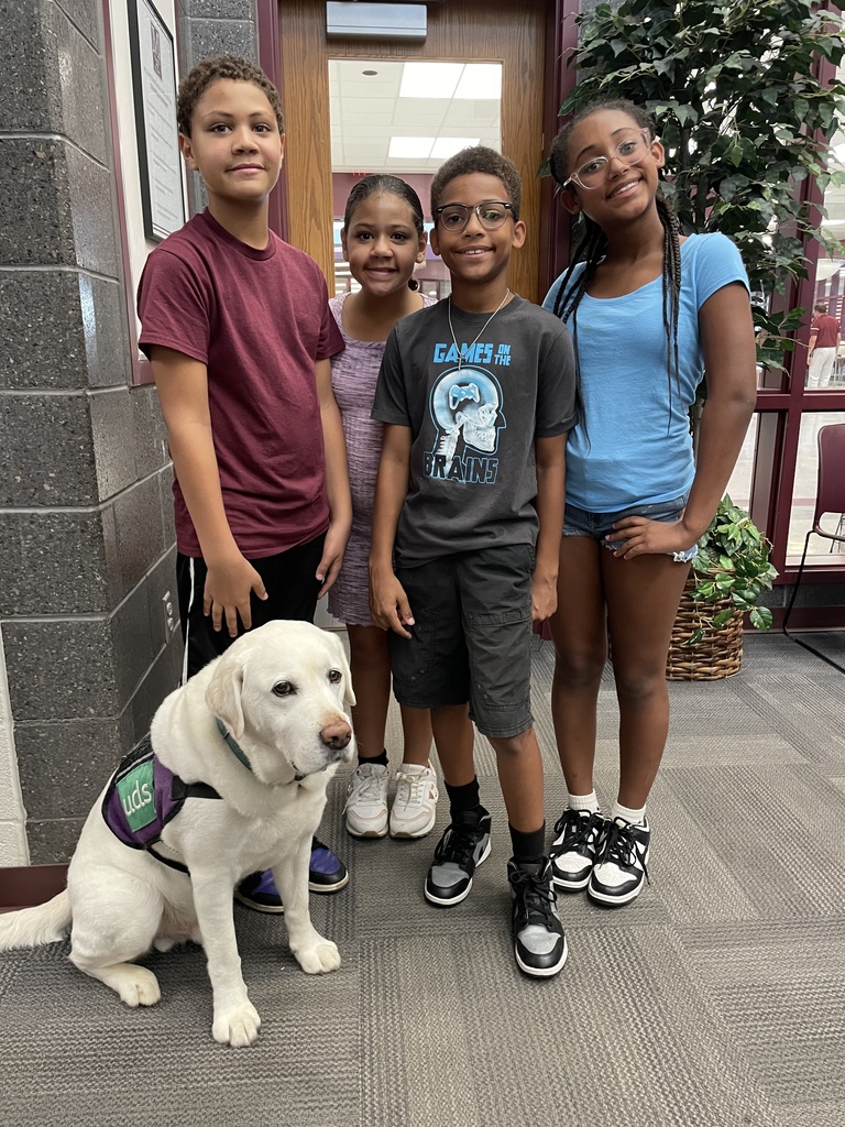 Four students standing with a white facility dog in a school hallway.
