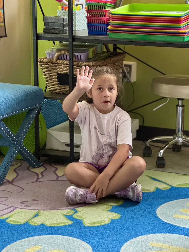 Little girl sitting criss cross on the floor with her hand raised.